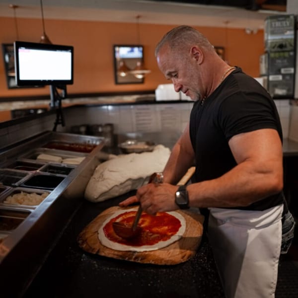 A chef at Esposito's Pizza Bar spreads red sauce on a pizza crust with a ladle.