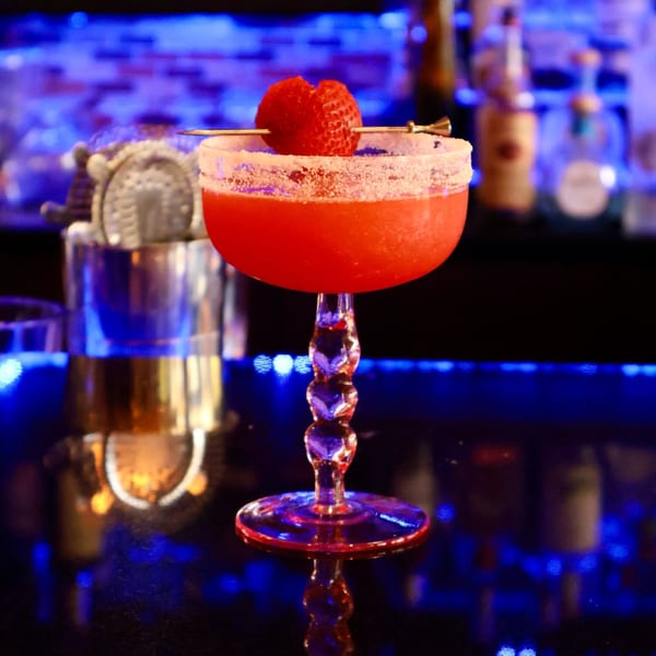 A vibrant red cocktail with a sugared rim and strawberry garnish sits on a bar with blue lighting.