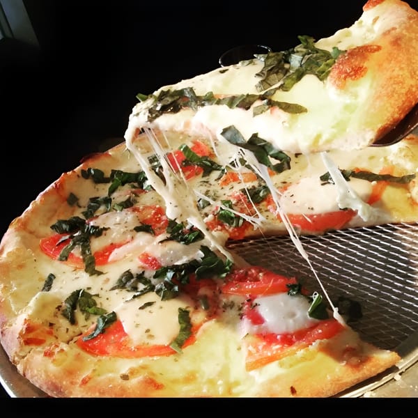 A slice of Esposito's Pizza Bar's Italian pizza with melted cheese, tomatoes, and basil being lifted.