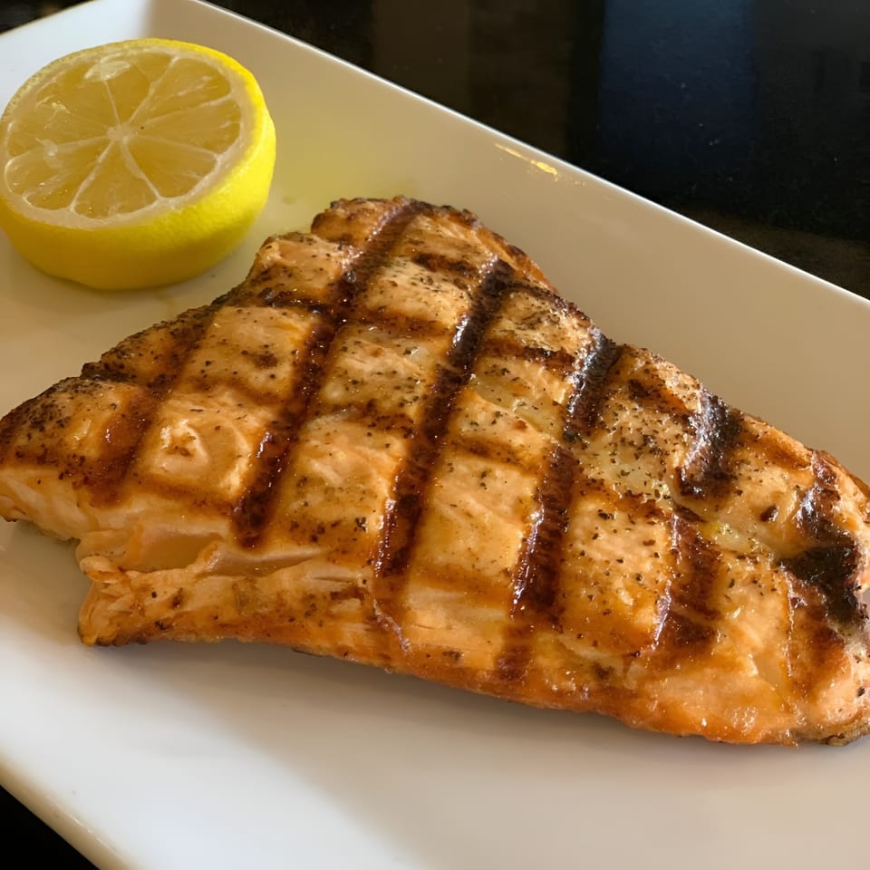Grilled salmon fillet with grill marks and a lemon half on a white rectangular plate.